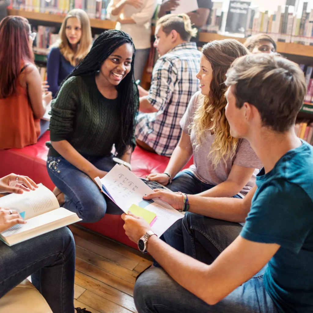 Foreign language tutoring session with diverse students studying together and discussing notes in a classroom library setting.